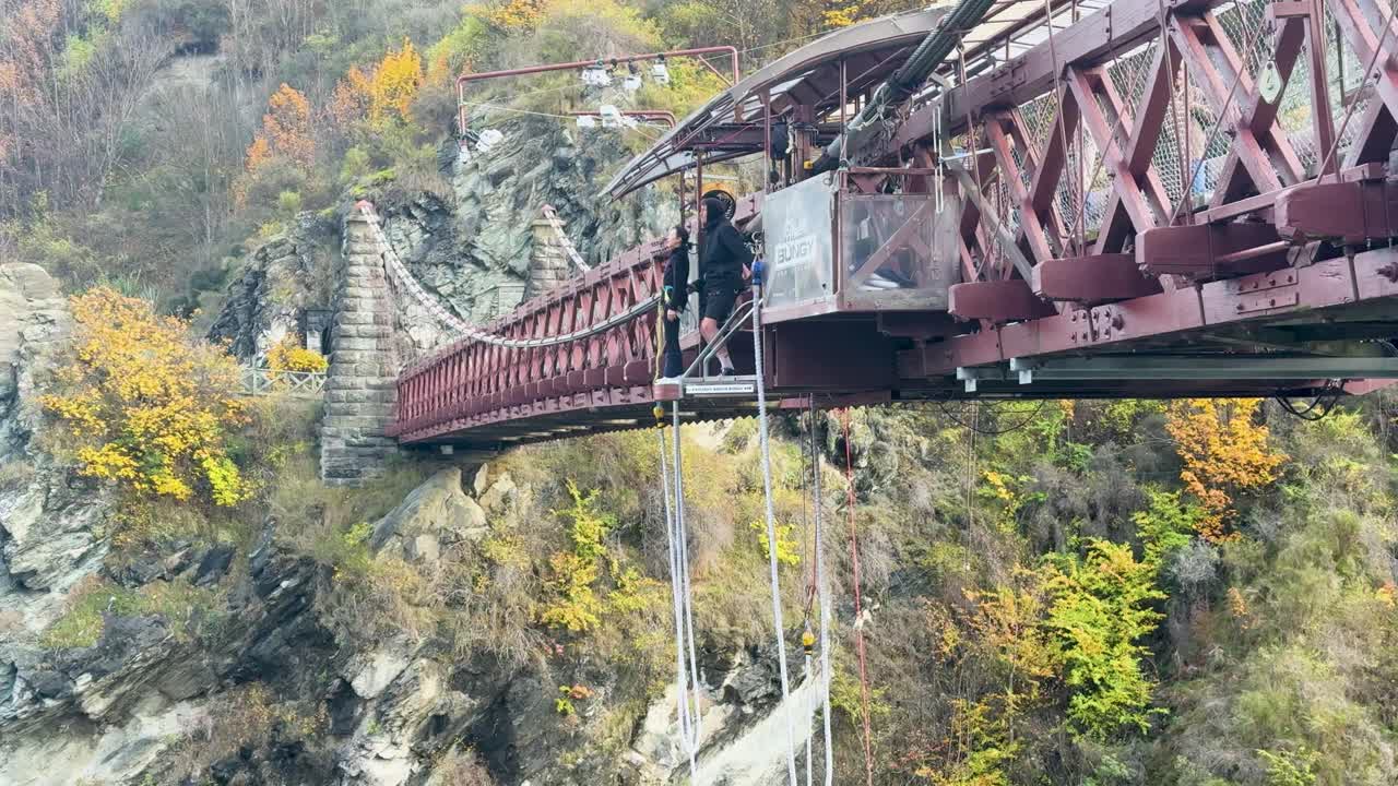 A person prepares for a bungee jump from a bridge in Queenstown, New Zealand, surrounded by stunning autumn foliage and rugged cliffs