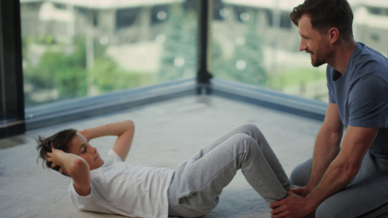 padre ayudando a su hijo en el entrenamiento en el interior. hijo haciendo ejercicios abdominales al lado de papá.