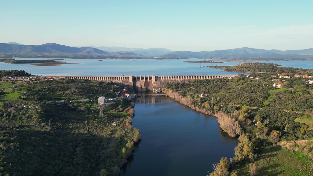 Vista aerea pulita con drone di una diga idroelettrica con bacino idrico e fiume, circondata da montagne e paesaggio rurale al tramonto