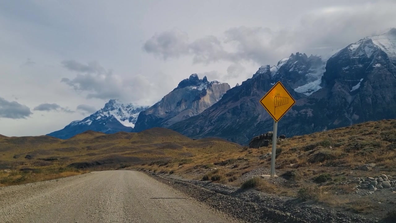 Keltainen tienvarsivaroitusmerkki voimakkaista tuulista Torres del Painen kansallispuistossa, Chilessä. Karut Patagonian vuoret, dramaattinen erämaa ja laaja alppimaisema avaran taivaan alla.