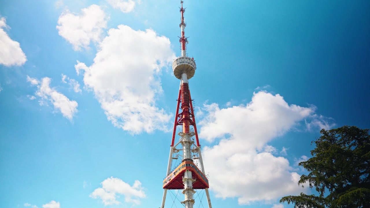 La torre televisiva rossa e bianca di Tbilisi si erge in un cielo azzurro sopra gli alberi e l'ingresso in vetro del Mtatsminda Park