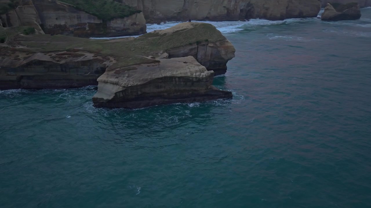 Aerial drone footage flying alongside the rugged limestone cliffs of Tunnel Beach at sunrise near Dunedin, New Zealand. Gentle ocean swells and soft morning light create a stunning coastal scene.