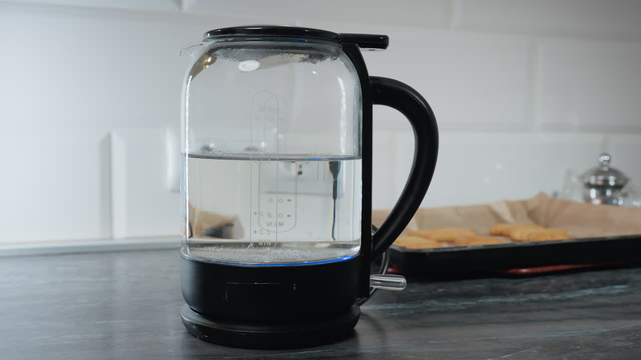 Close-up of person turning off electric water boiler after boiling, with water level shown in jar, background features kitchen with cookies, a kettle, and kitchen appliances
