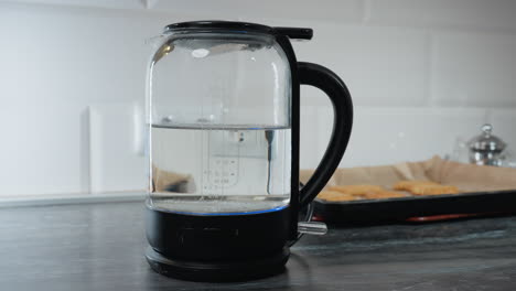 primer plano de una persona apagando la caldera de agua eléctrica después de la ebullición, con el nivel de agua mostrado en el frasco, el fondo presenta una cocina con galletas, un hervidor y aparatos de cocina