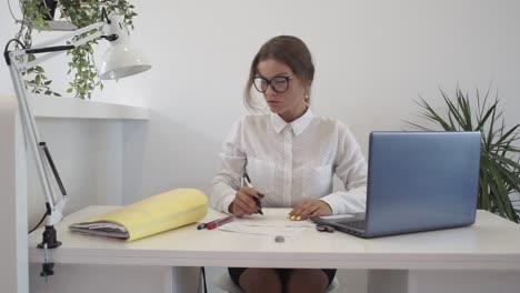 a woman at the office fills out some paperwork, glances out the window before getting an idea and then starts typing on her laptop