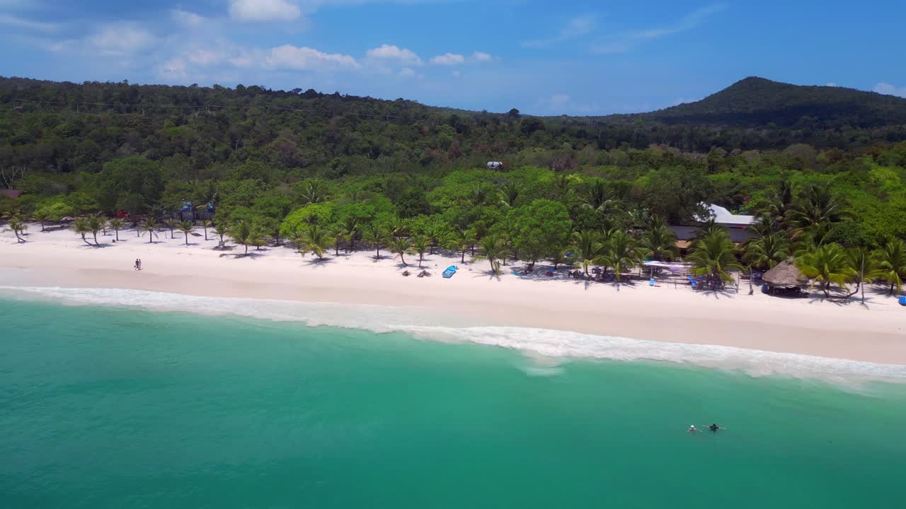 Long Set Beach in Koh Rong island, featuring turquoise waters, blue boat on sand beach and lush tropical vegetation. Amazing aerial view flight fly reverse drone