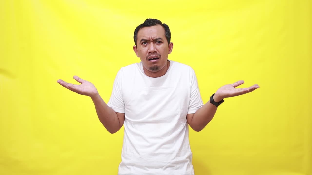 Asian man shrugs shoulders unaware raises hand clueless has no idea cannot answer poses against yellow background