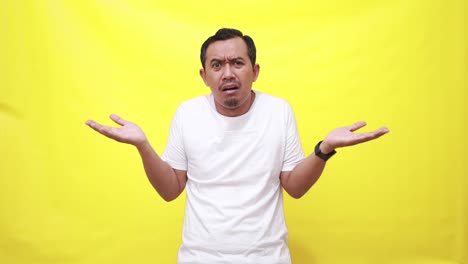 Asian-man-shrugs-shoulders-unaware-raises-hand-clueless-has-no-idea-cannot-answer-poses-against-yellow-background