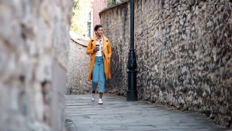 young black woman wearing an unbuttoned yellow pea coat and jeans walking in an alley in historical district listening to music and holding a takeaway coffee, low angle