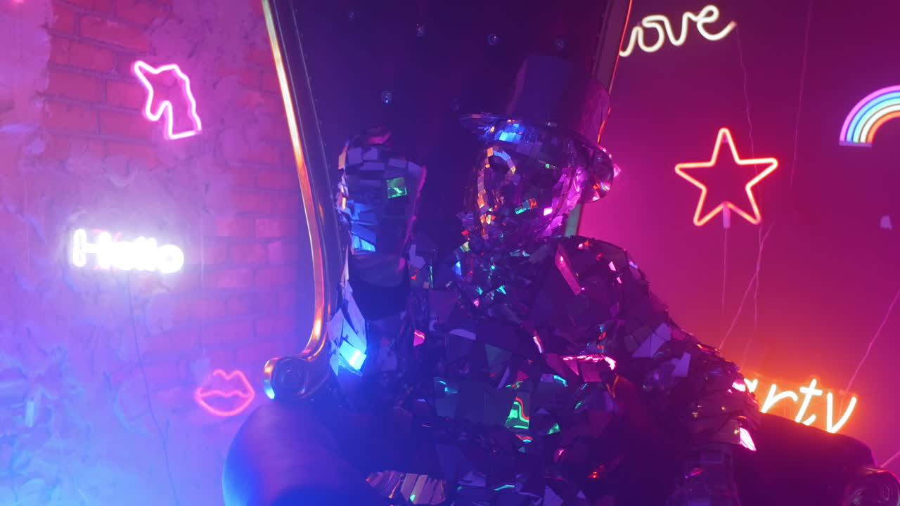 un hombre con un traje de metal plateado de acero se sienta en una silla y sacude la cabeza a la música en la luz de neón. dedo se hace señas a sí mismo. llamar a sí mismo con un gesto.