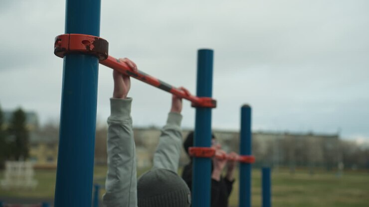 Two boys are working out on pull-up bars in an outdoor gym area, the boy in the gray sweater struggles to continue, while the boy in the black sweater pauses and looks at him