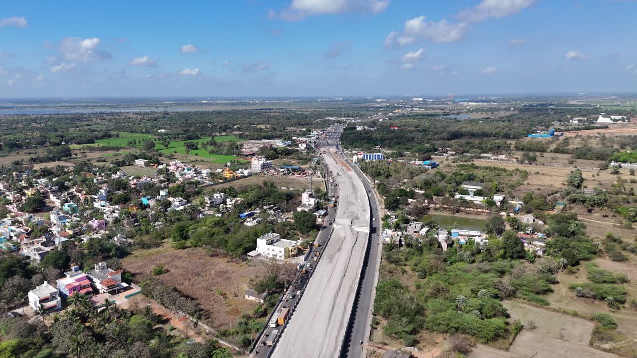 A major highway construction project near Chennai, with new sections and traffic on existing roads. Residential areas, fields, and a large lake are visible, urban growth under a partly cloudy sky