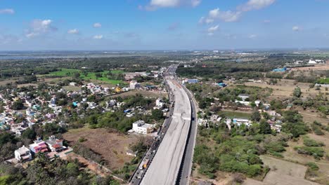 a major highway construction project near chennai, with new sections and traffic on existing roads. residential areas, fields, and a large lake are visible, urban growth under a partly cloudy sky