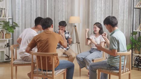 아시안 십대 그룹은 의자에 앉아서 집에서 스마트폰으로 게임을 하는 원을 형성합니다. 성공을 축하하고 게임 경기에서 승리하고, 하이 파이브 제스처를 합니다.