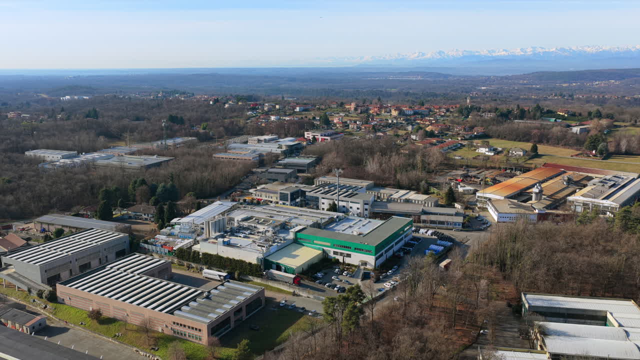 Aerial drone view shows an industrial area in northern Italy featuring manufacturing plants, warehouse roofs, internal road networks, and surrounding countryside, Swiss Alps rising in background