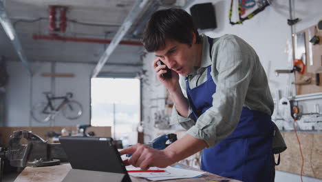 propietario de un negocio masculino en un taller usando una tableta digital y haciendo llamadas por teléfono móvil