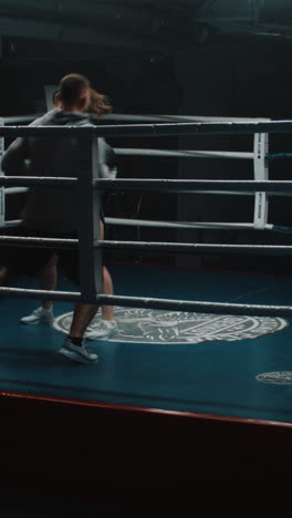 athletic woman in boxing gloves prepares for fighting competition and exercises with male trainer in dark gym. female boxer hits punching mitts on boxing ring. vertical shot