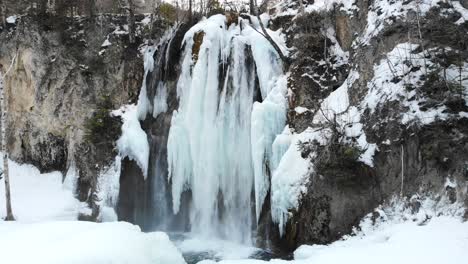 Rising-shot-above-partially-frozen-waterfall-to-reveal-mountains-and-forest-in-winter-with-snow-in-4K