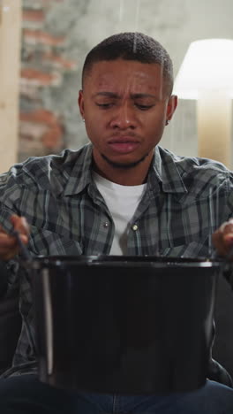 disappointed black man collects water into basin during leakage from upper floor in living room. pipe break causes apartment flooding. plumber service