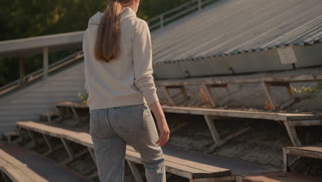 back view of young woman in casual hoodie and jeans walking towards bleachers in outdoor stadium, soft sunlight highlights her relaxed posture as she approaches rustic seating area