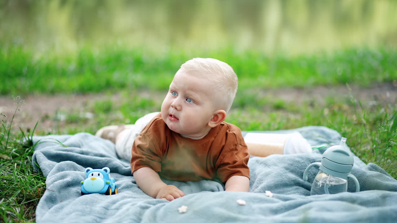 Lovely blond toddler kid lies outdoors on the plaid. Baby boy puts a flower in the mouth and chews it carefully.