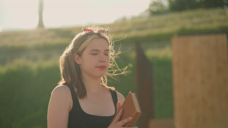 woman closes the book in her hand, takes a deep breath, as wind blows her hair, the sun radiates on her face, and she gazes into the distance, with wooden structure in the background