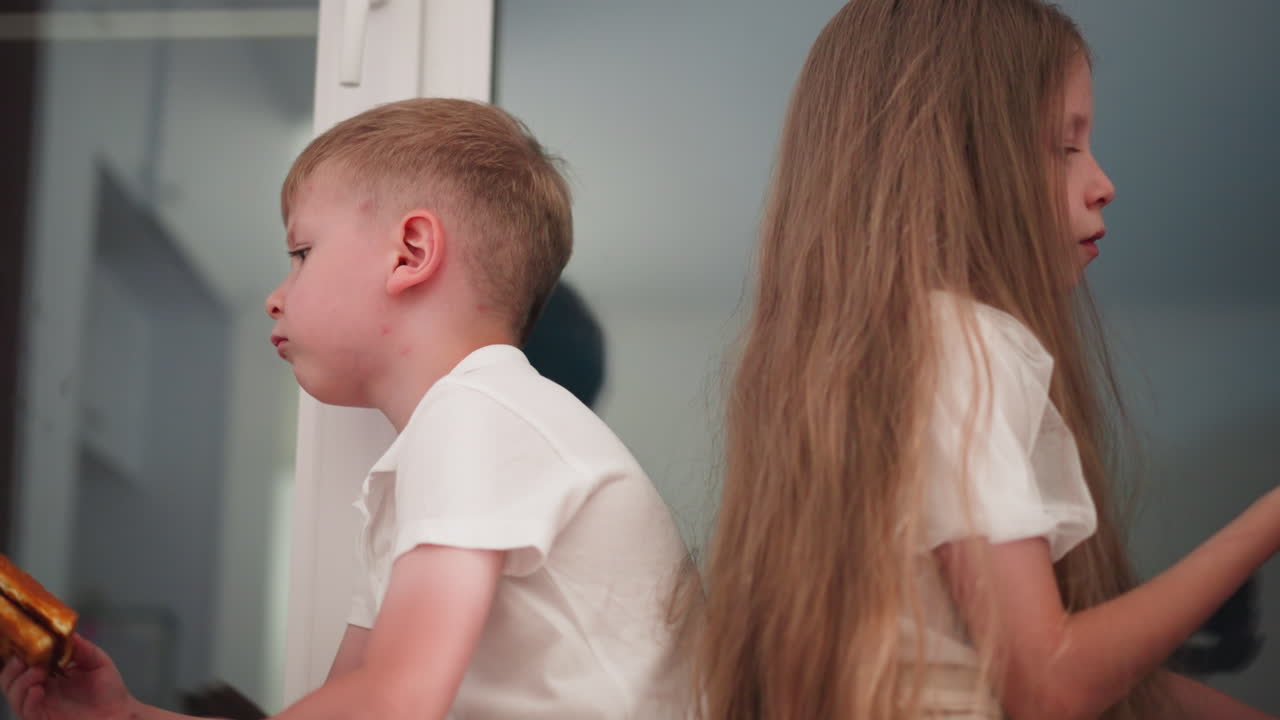 Siblings seated with their backs resting on each other, enjoying dessert while eating, creating a peaceful and relaxed moment together, with a subtle reflection visible in the glass behind