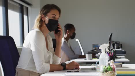 two diverse female colleagues wearing face mask, working at desk in office