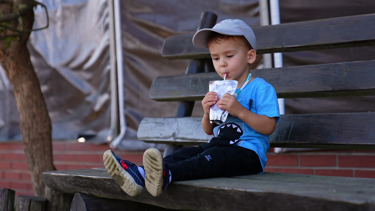 Calm Caucasian baby boy sitting on the bench outdoors drinking juice from doy pack. Thirsty child drinking with straw.
