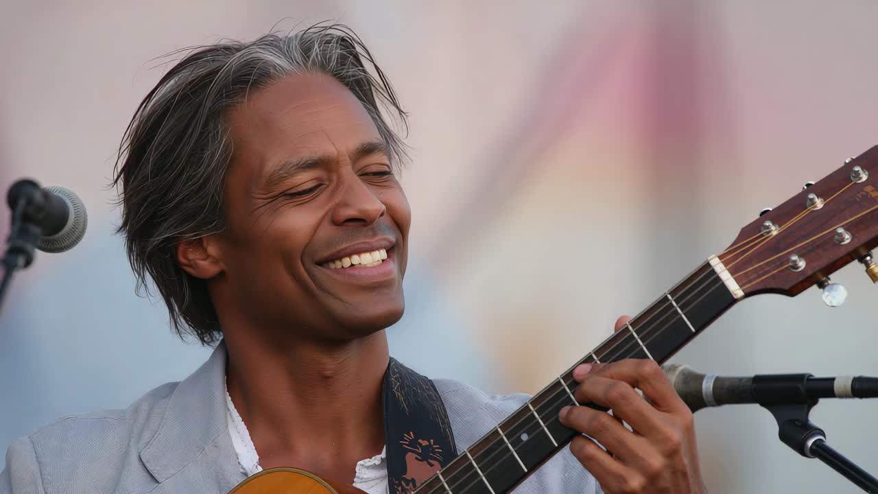 Sorride, guarda la tastiera, musicista in blazer grigio che suona la chitarra acustica al microfono sul palco