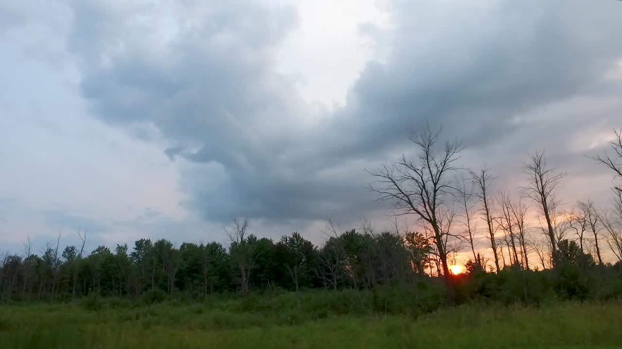 Time lapse driving alongside wooded area with sun setting in distance.