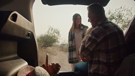 A-happy-man-with-gray-hair-in-a-checkered-shirt-meets-his-wife,-a-brunette-girl-in-a-green-checkered-shirt,-and-their-little-daughter-running-towards-him-while-the-man-sits-in-the-open-trunk-of-a-car-and-looks-at-his-loved-ones-during-a-picnic-in-the-summer