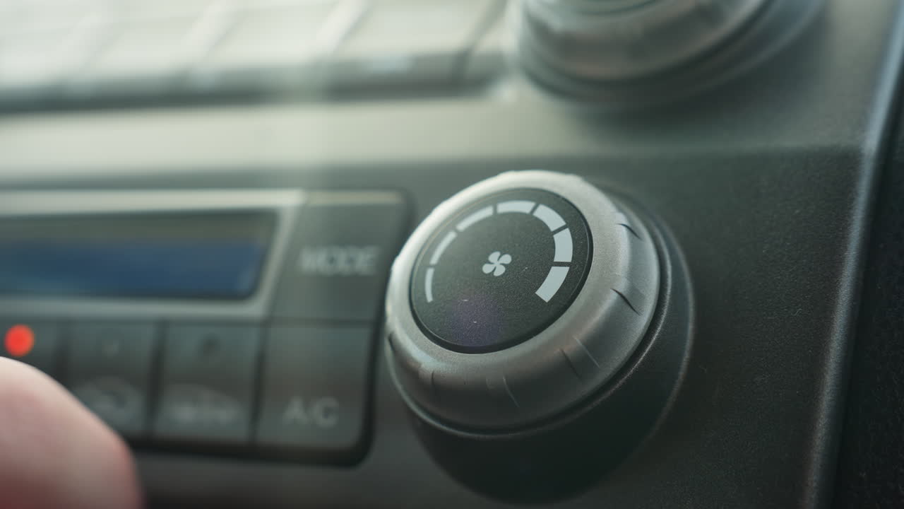 close up hand rotating ac control knob inside car dashboard climate control panel showing white fan icon segments around knob ambient sunlight glare reflection