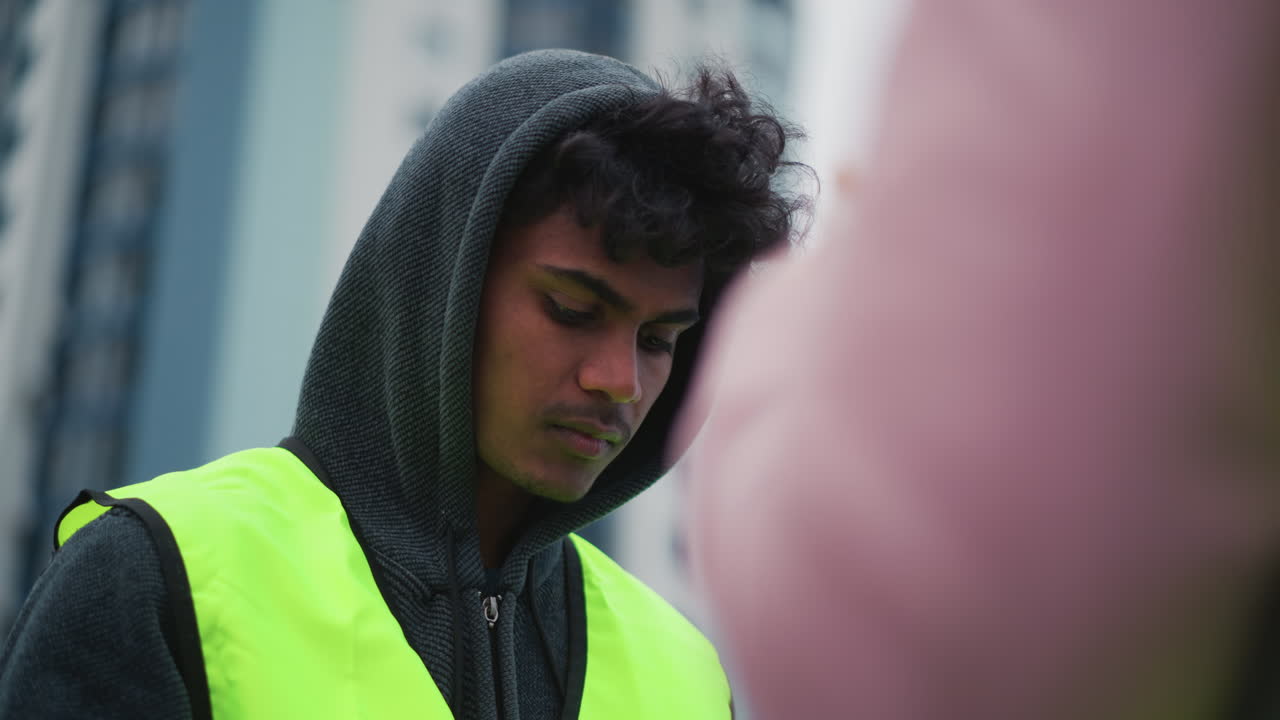 Young male worker in dark hoodie and neon reflective vest standing outdoors near modern city buildings, looking serious and attentive during work, with blurred person in foreground urban environment