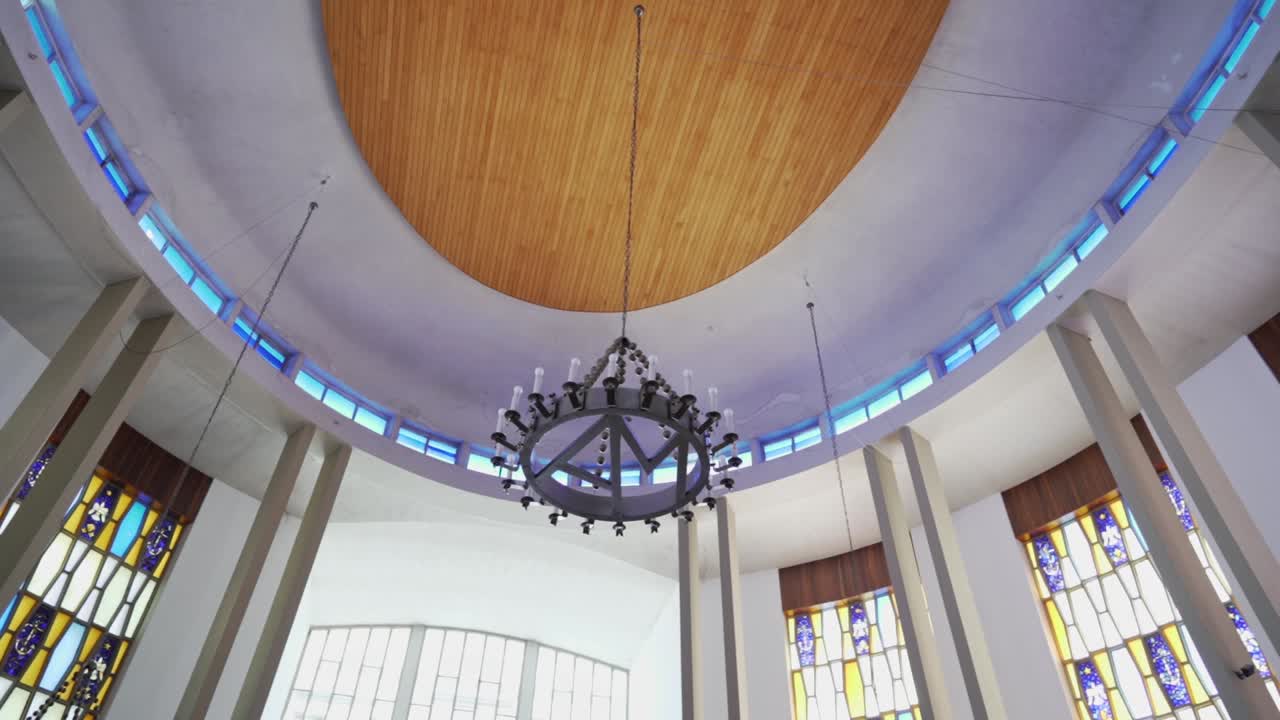 Vista verso l'alto della cupola interna di una chiesa con lampadario circolare, alte colonne e vetrate colorate illuminate da una morbida luce diurna.
