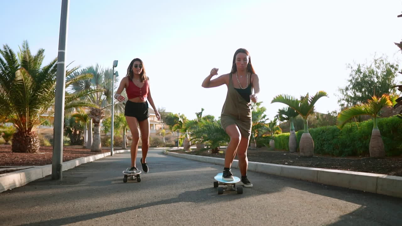 Two young Hispanic women ride skateboards on an island near palm trees, cruising on the park's asphalt paths in slow motion under the sunset's warm hues. Happiness and a healthy lifestyle