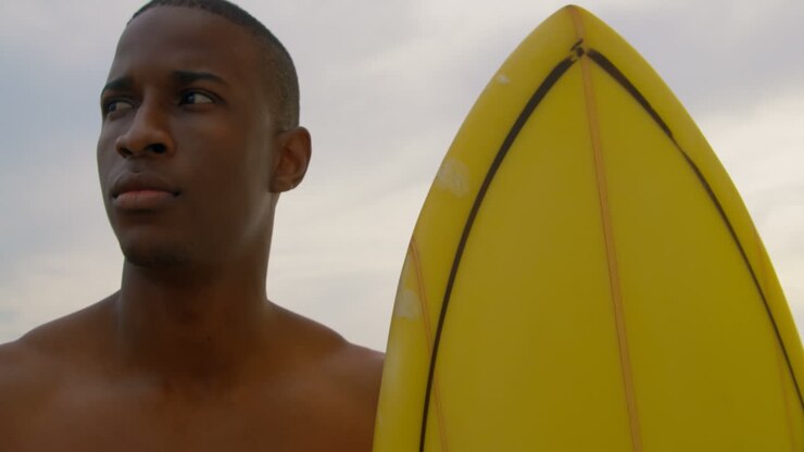 Vista frontale di un surfista afroamericano in piedi con la tavola da surf sulla spiaggia 4K.