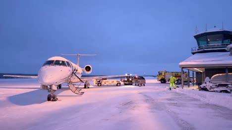 a jet sits on a snowy runway as airport crew and vehicles prepare it under the twilight skies of enontekiö, finland - medium shot