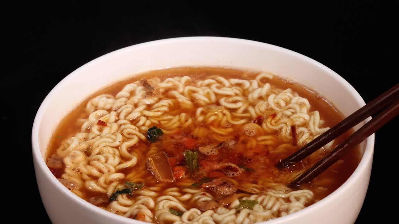 Chopsticks gently stir a bowl of instant noodles in broth under even studio lighting, with a static overhead camera capturing the motion against a black background