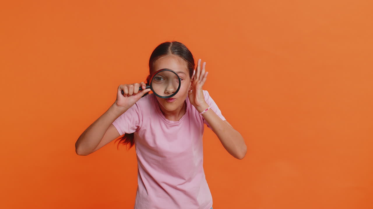 Child girl kid holds magnifying glass near face looking at camera with big zoomed eye analyzing