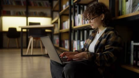 Happy-girl-student-with-curly-hair-sits-near-shelves-with-books-and-types-on-a-laptop-in-the-library