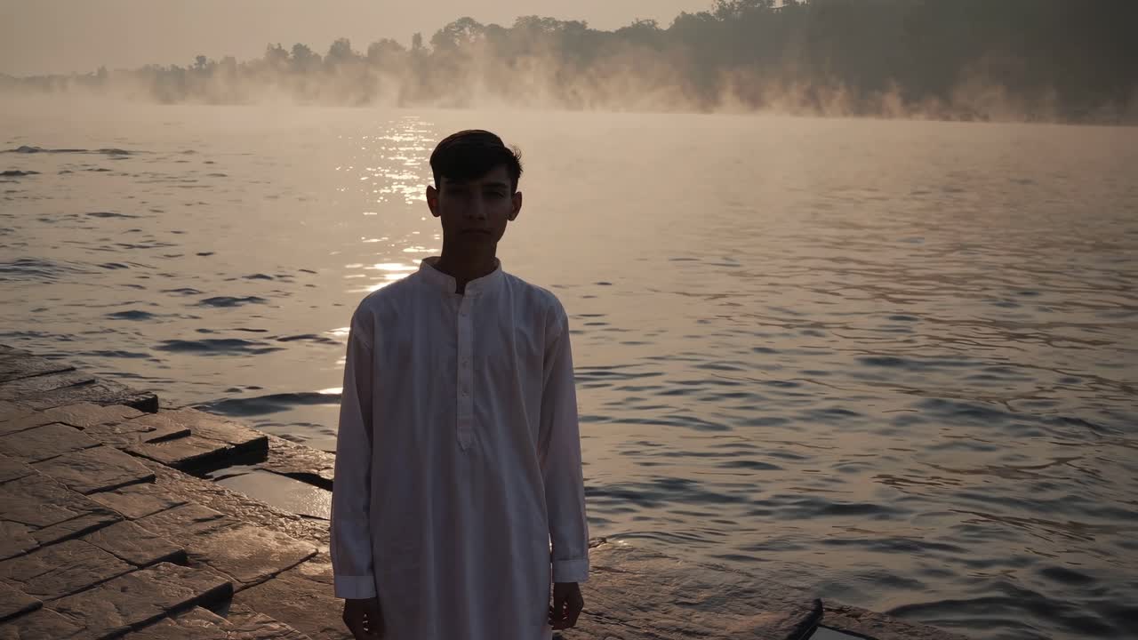 Young male figure in white attire stands by serene riverbank at dawn, with mist rising from water and soft sunlight reflecting on surface, capturing tranquil moment of contemplation