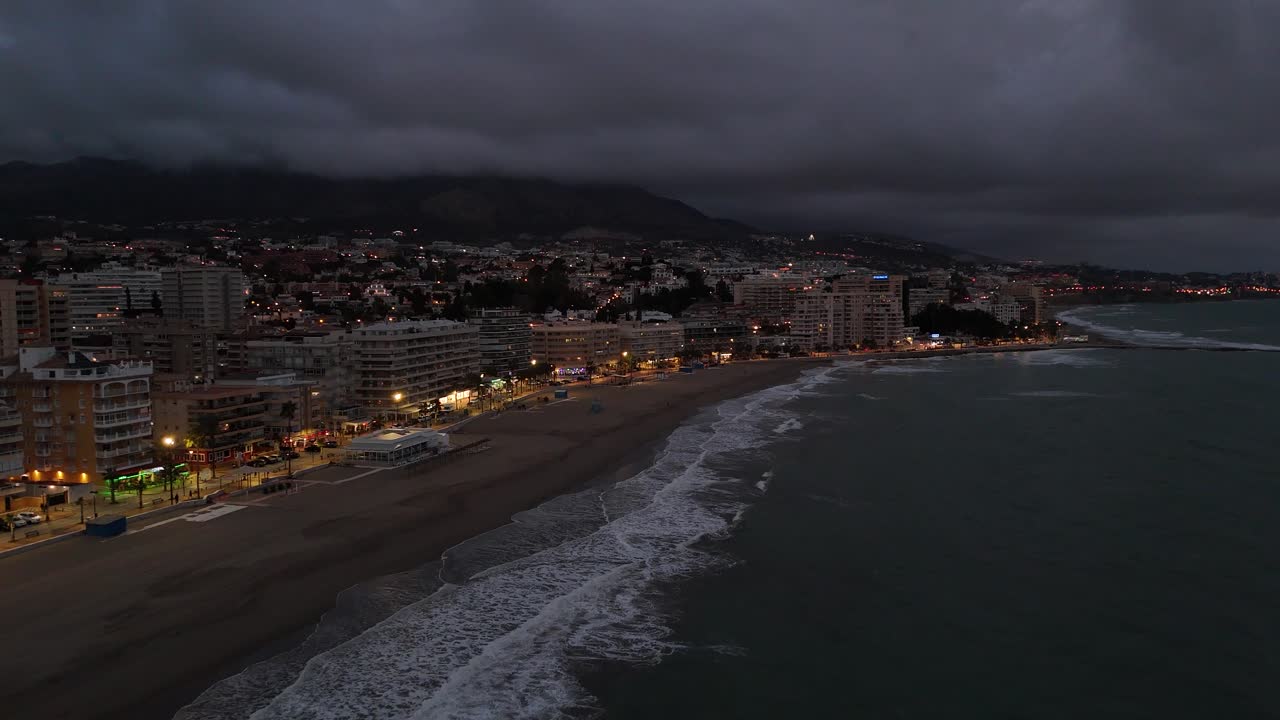 Aerial view of Torreblanca, Spain at dusk, showing illuminated buildings along the coastline, waves crashing on the beach, and dark clouds covering the mountains