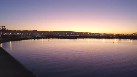 Großflächiger-Schifffahrtshafen-Mit-Bergkulisse-Und-Strahlend-Blühendem-Sonnenaufgang-über-Purple-Bay