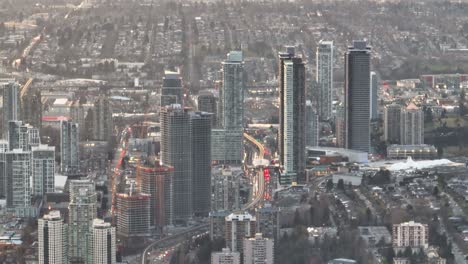 Aerial-truck-left-of-highway-traffic,-Sky-Train-and-Brentwood-Town-Centre-skyscrapers-in-Burnaby-city-skyline-at-daytime,-British-Columbia,-Canada