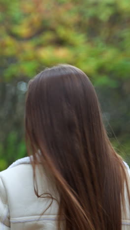 smiling brunette in light warm jacket walking in the nature. girl taking a stroll in the park in autumn. blurred backdrop. vertical video