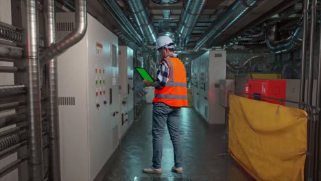 full body back view of asian male engineer with safety helmet working on a green screen tablet and looking around while standing in engine control room, work of electrical generators
