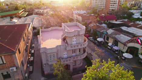 Flyover-of-the-Sermini-Palace-or-Castillito-de-los-Jesuitas-in-Providencia-Santiago-with-trees-illuminated-by-a-beautiful-orange-sunset-light,-pedestrians-walking-along-the-signage