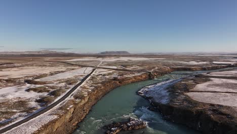 aerial view of urriðafoss waterfall and surrounding winter landscape in iceland, featuring icy riverbanks, winding roads, and a vast open terrain under a clear blue sky.