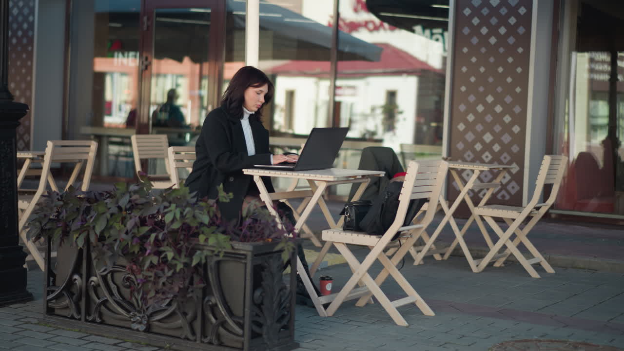 mujer trabajando en una computadora portátil al aire libre en un restaurante, escribiendo en una mesa con plantas y una taza de café cerca, centrada en la tarea, con un fondo borroso